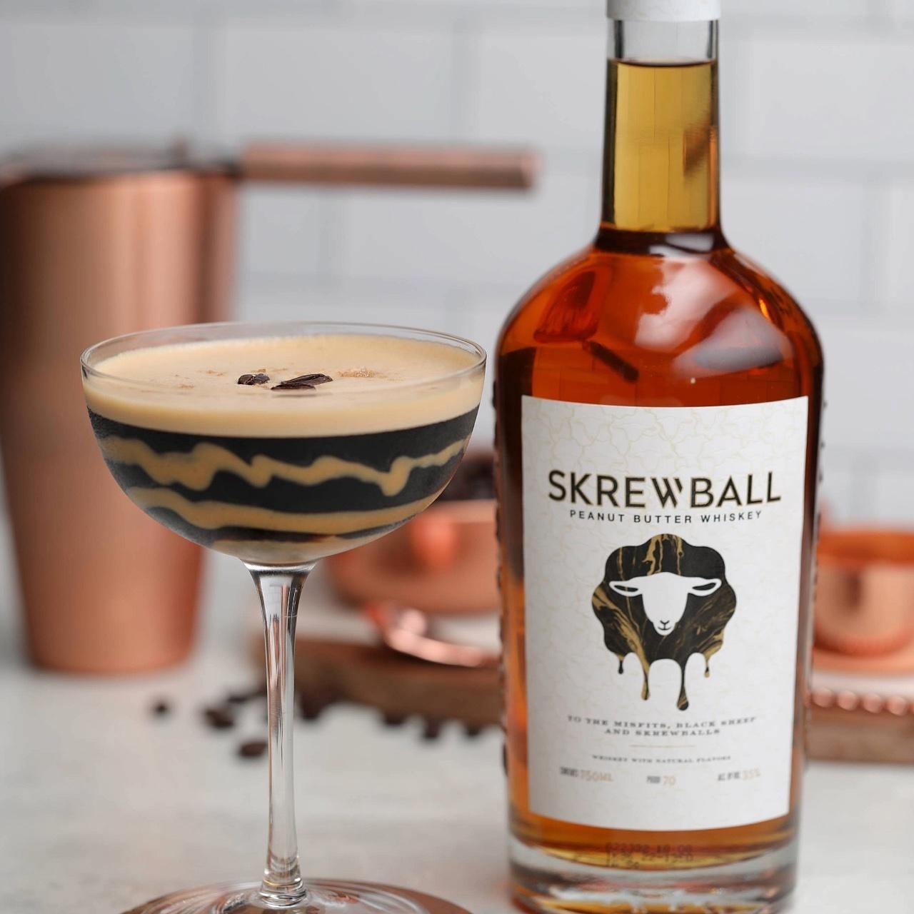 A bottle of Skrewball Peanut Butter Whiskey next to a cocktail glass filled with a drink topped with coffee beans, set on a kitchen counter