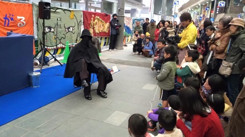 観客の前で布を被った人がパフォーマンスを行っています。多くの子供や大人が興味深く見つめています。