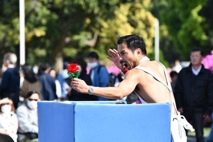 男性が群衆の前でステージ上に立ち、赤と緑の花を持っています。背景に多くの人々がいます。