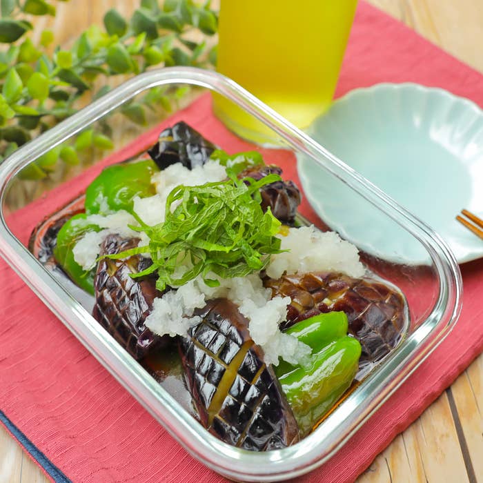 透明な容器に盛られた焼きナスとピーマン、おろし大根とシソの葉が上に乗っています。鮮やかな飲み物が背景にあります。