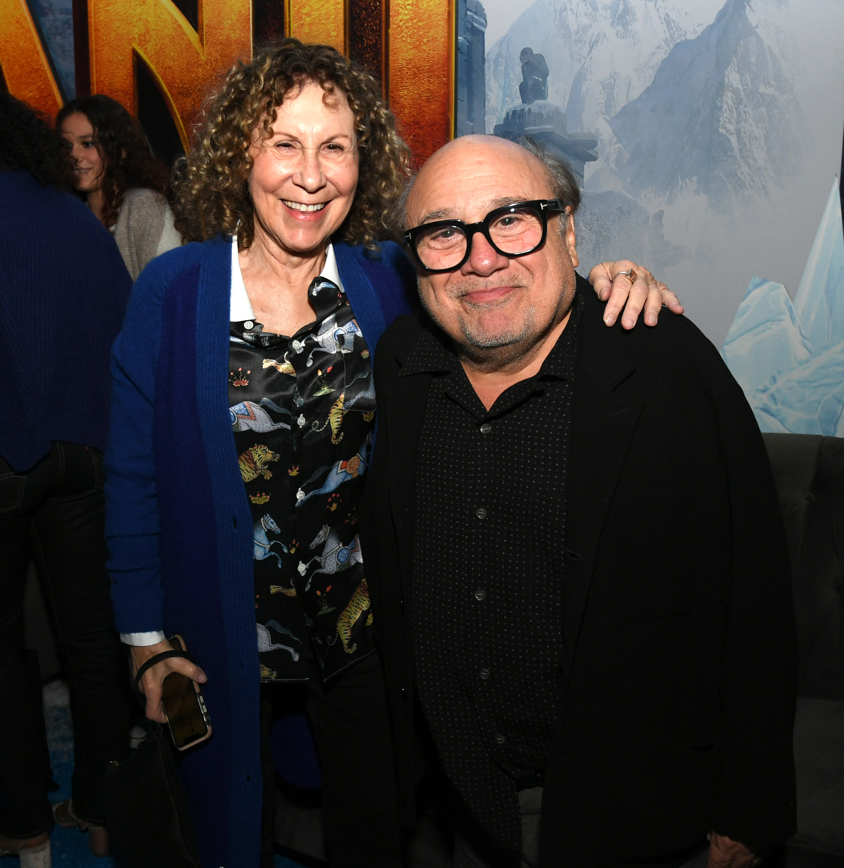 Rhea Perlman and Danny DeVito are smiling and standing next to each other at an event. Rhea is wearing a patterned shirt with a cardigan, and Danny is wearing a dark shirt with glasses