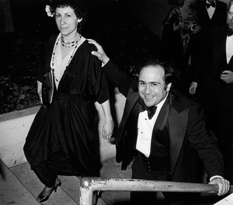 Danny DeVito, in a tuxedo, and Rhea Perlman, in a dark dress, smile while climbing stairs at an event