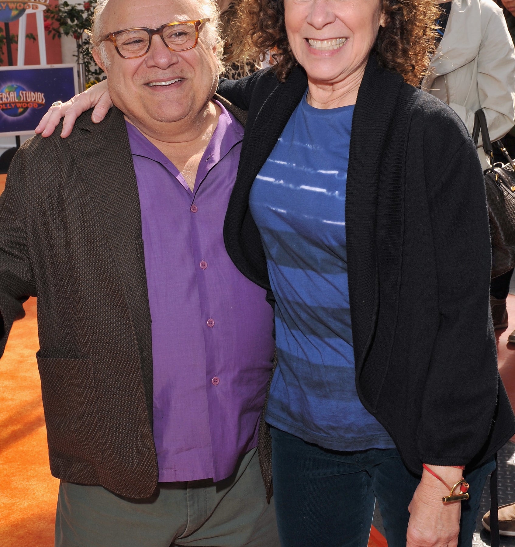 Danny DeVito in a blazer and open-collar shirt, and Rhea Perlman in a casual top and cardigan, smile together at a red carpet event