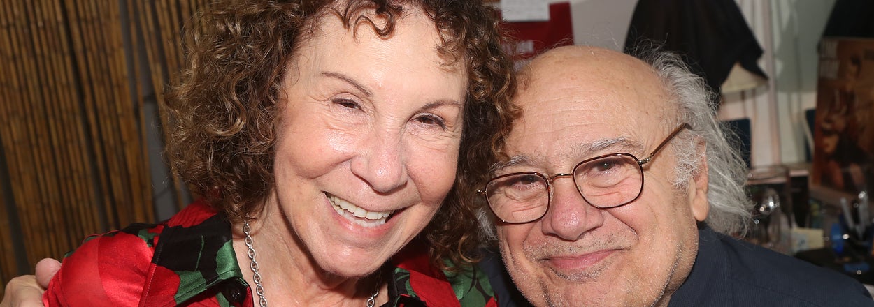 Rhea Perlman and Danny DeVito smiling together, with Rhea wearing a floral patterned shirt and Danny in a casual button-up