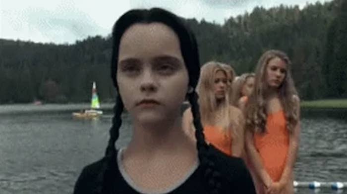 Scene from The Addams Family featuring Wednesday Addams with long dark braids standing in front of a lake, with girls in swimsuits behind her