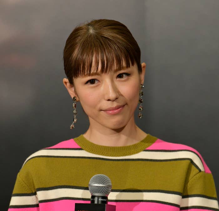 女性がマイクを持っている。彼女はボーダー柄のトップを着て、大きなイヤリングを身につけている。