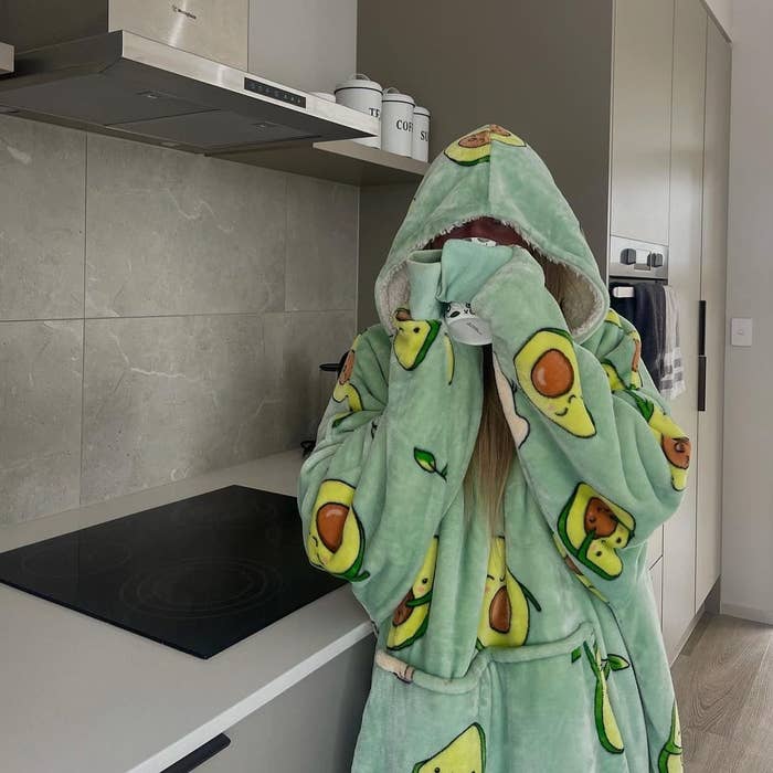 Person in an avocado-patterned robe hides their face with the robe hood in a modern kitchen. Identifiable names not provided