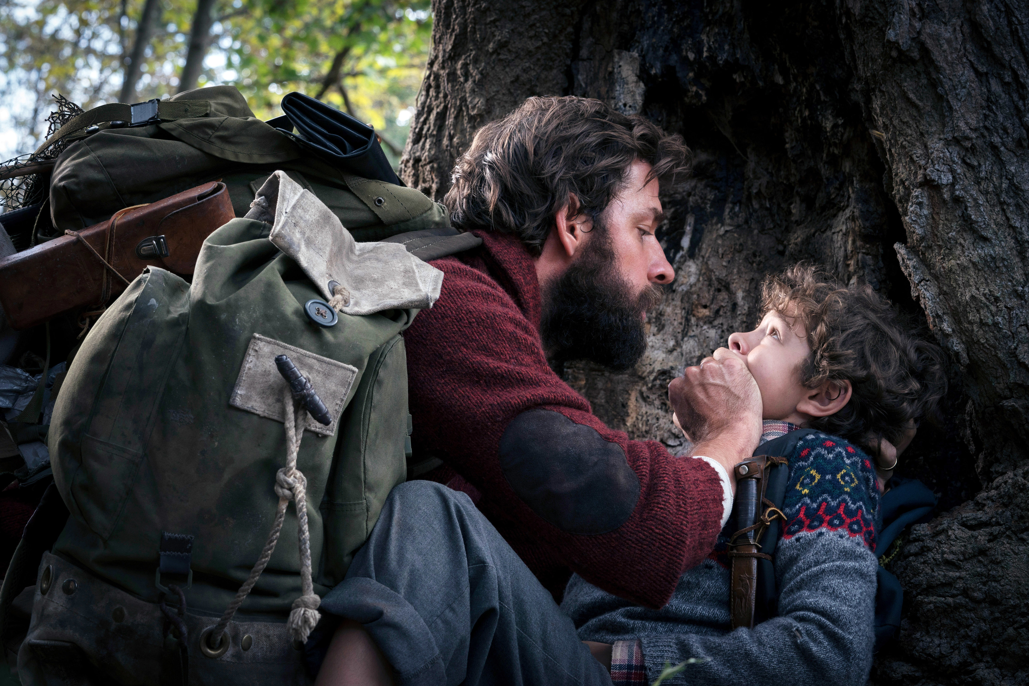 John Krasinski, in rugged clothes, hushes a fearful Noah Jupe against a tree in a tense outdoor scene