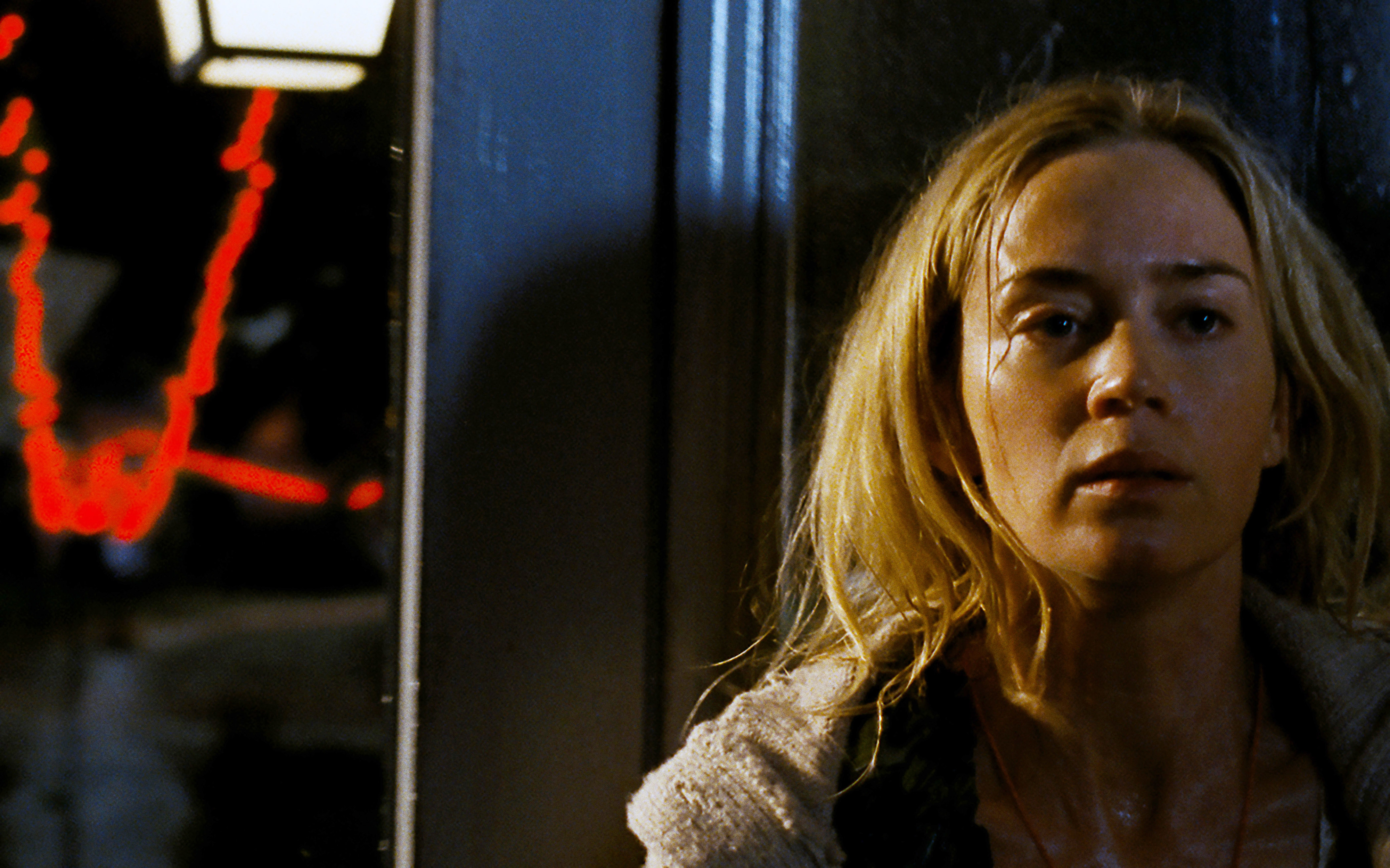 Emily Blunt, with disheveled hair and a concerned expression, leans against a door in a dimly lit scene