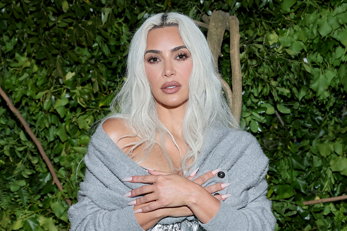 Kim Kardashian with platinum blonde hair, wearing an off-the-shoulder gray cardigan over a silver top, posing in front of foliage