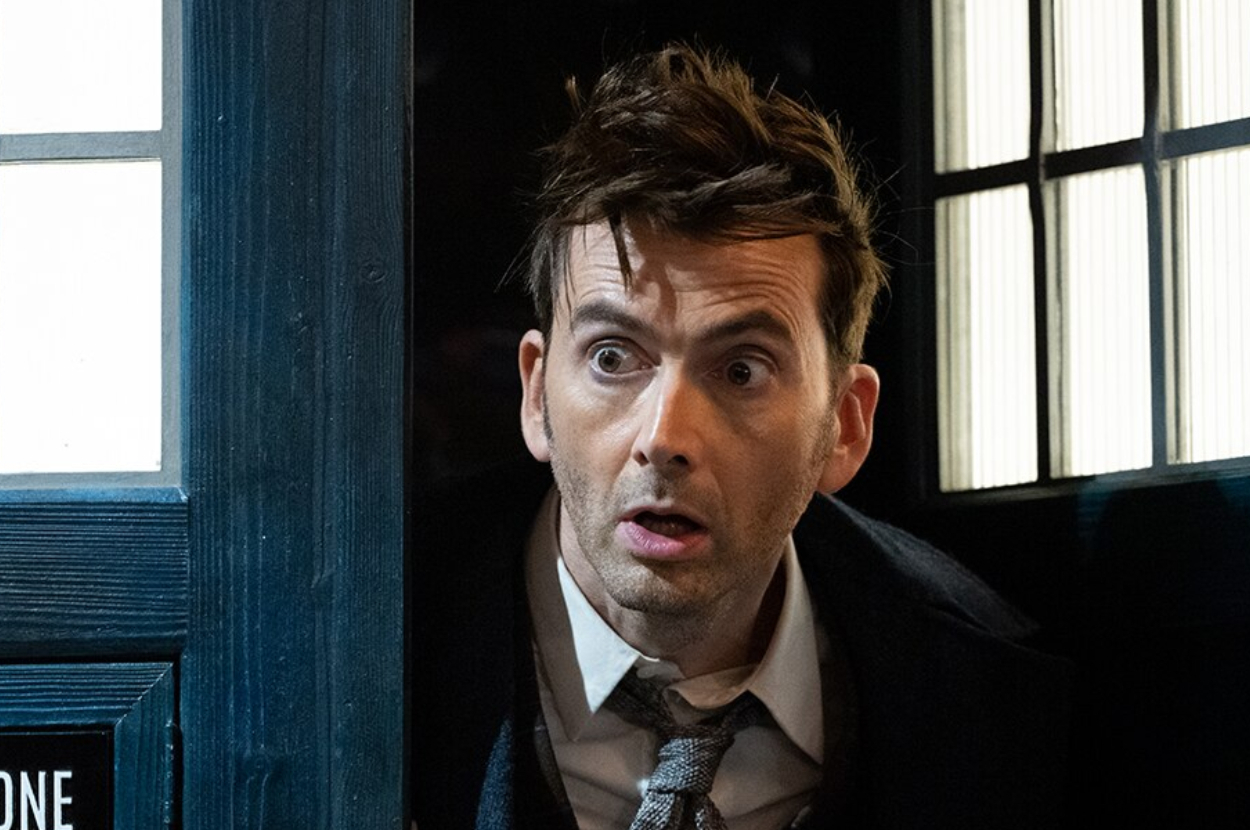 David Tennant, dressed as the Doctor in a suit and tie, looks surprised while peering out of the TARDIS door