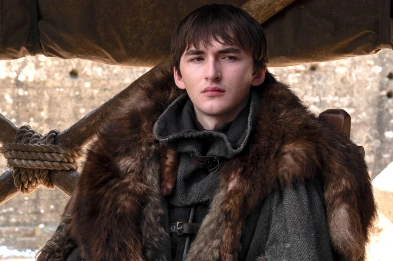 Isaac Hempstead Wright, wearing a medieval-style cloak with fur, stares into the distance in an outdoor setting