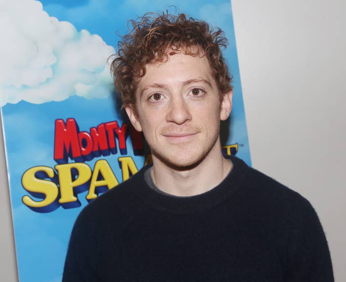 Josh Grisetti stands in front of a "Monty Python's Spamalot" promotional poster, wearing a casual dark sweater