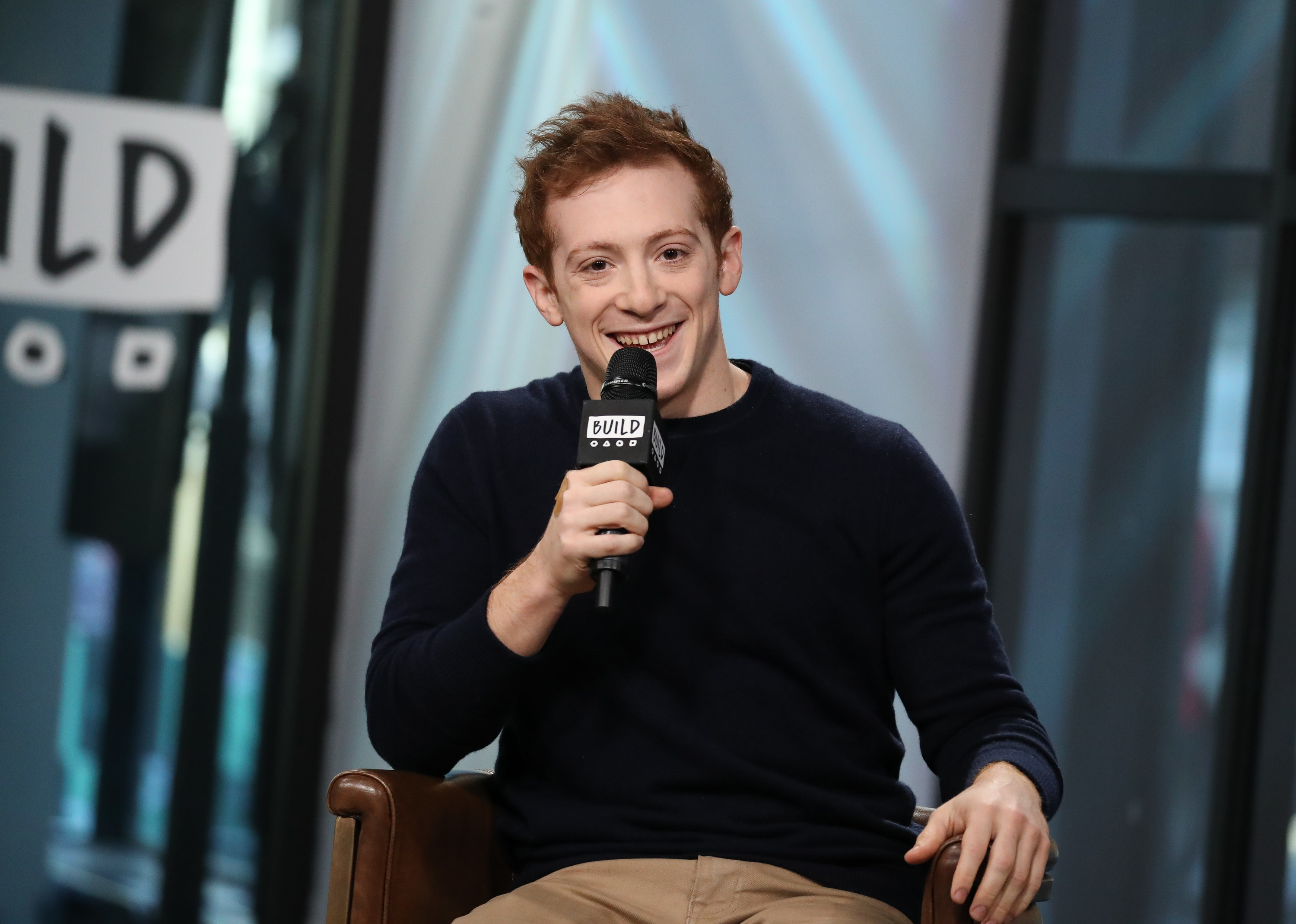 Ethan is seated, smiling, and holding a microphone during an interview