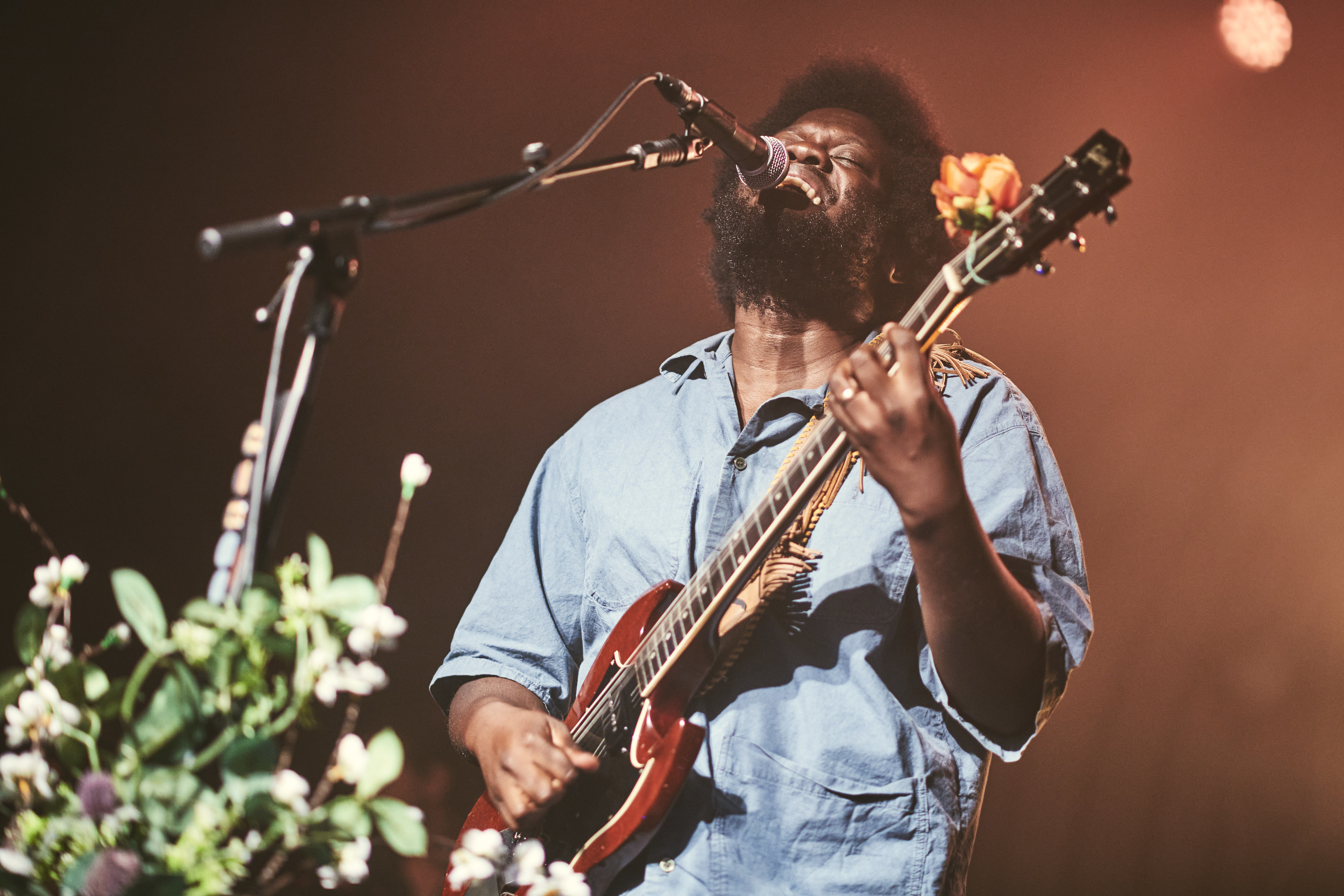 Michael Kiwanuka performs passionately on stage, singing into a microphone while playing an electric guitar adorned with a flower