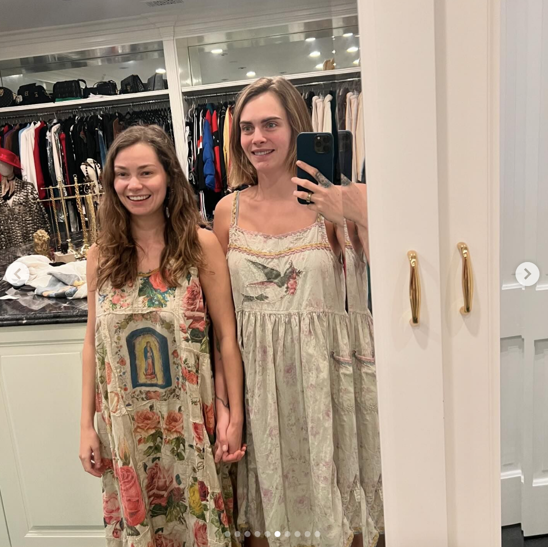 Minke and Cara Delevingne are standing and holding hands in a closet, both wearing flowy dresses with floral patterns, looking at the camera