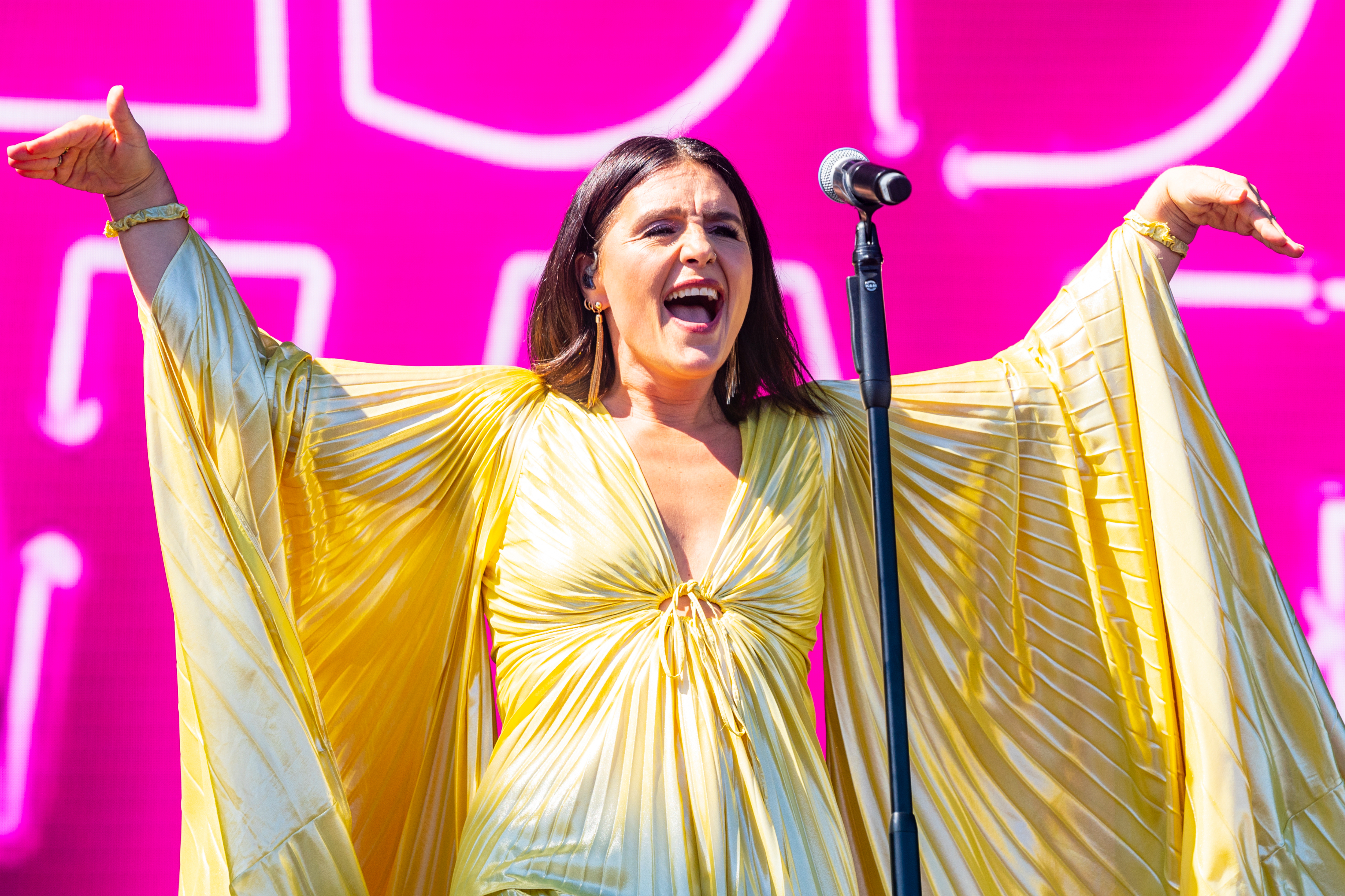 Jessie Ware performing on stage, wearing a flowing, pleated outfit with wide sleeves. She is smiling and raising her arms near a microphone
