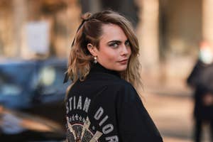 Cara Delevingne wearing a black Christian Dior jacket outdoors, looking over her shoulder at the camera, with an unfocused person in the background