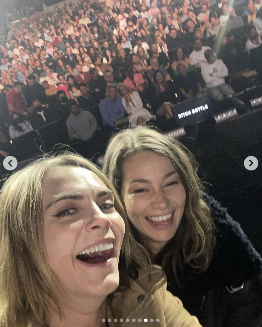 Cara Delevingne and a Minke take a selfie with a large audience in the background. Both are smiling broadly