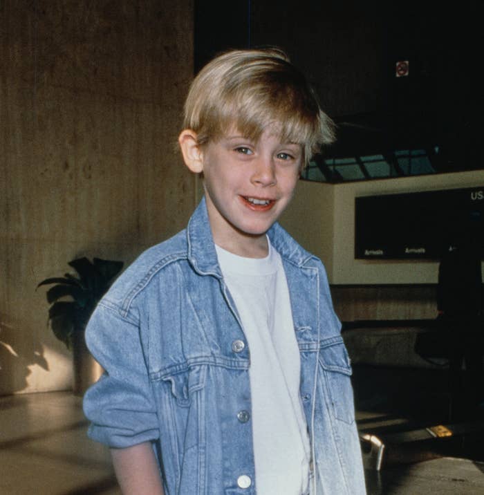 Macaulay Culkin, as a child, stands indoors wearing a light denim jacket and jeans with hands in his pockets. He smiles at the camera