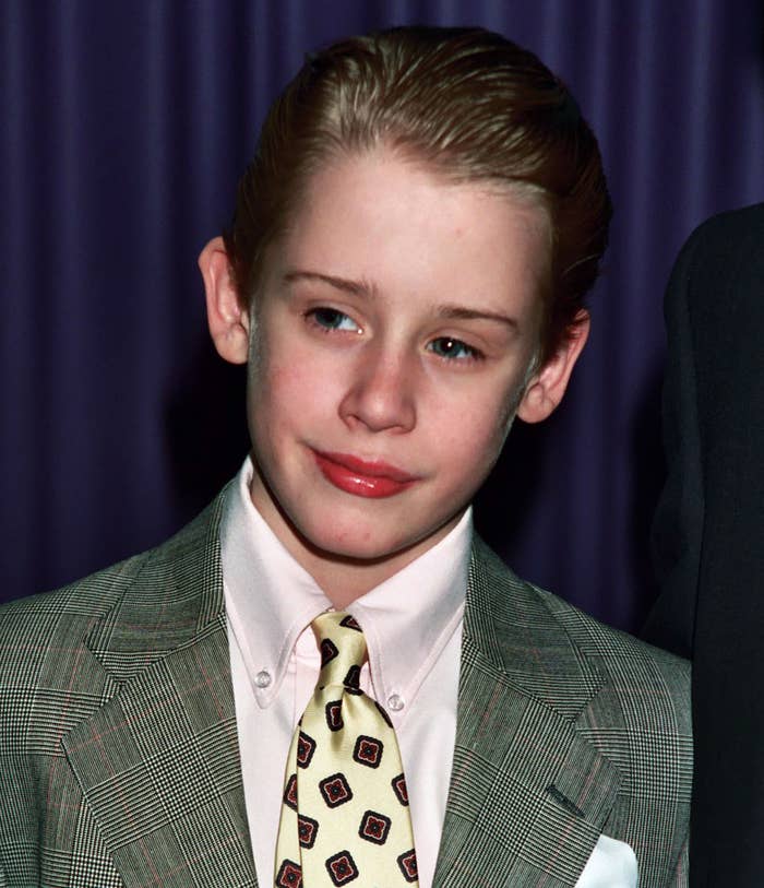 Macaulay Culkin, as a child, in a checkered suit and a patterned tie, poses in front of a curtain at an event