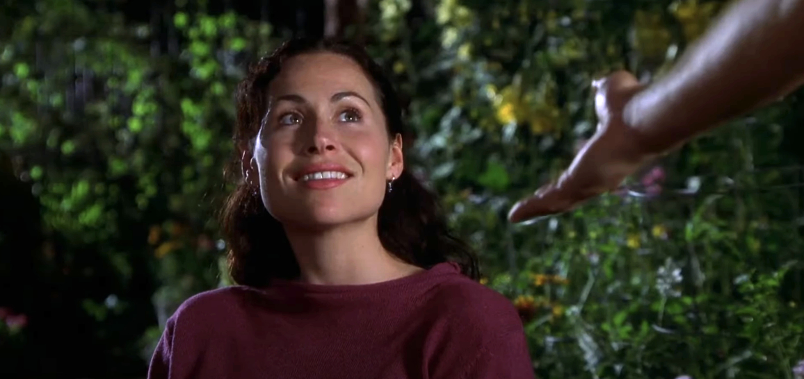 A person with dark curly hair looks up with a smile as an outstretched hand appears from the right side of the frame in a garden setting