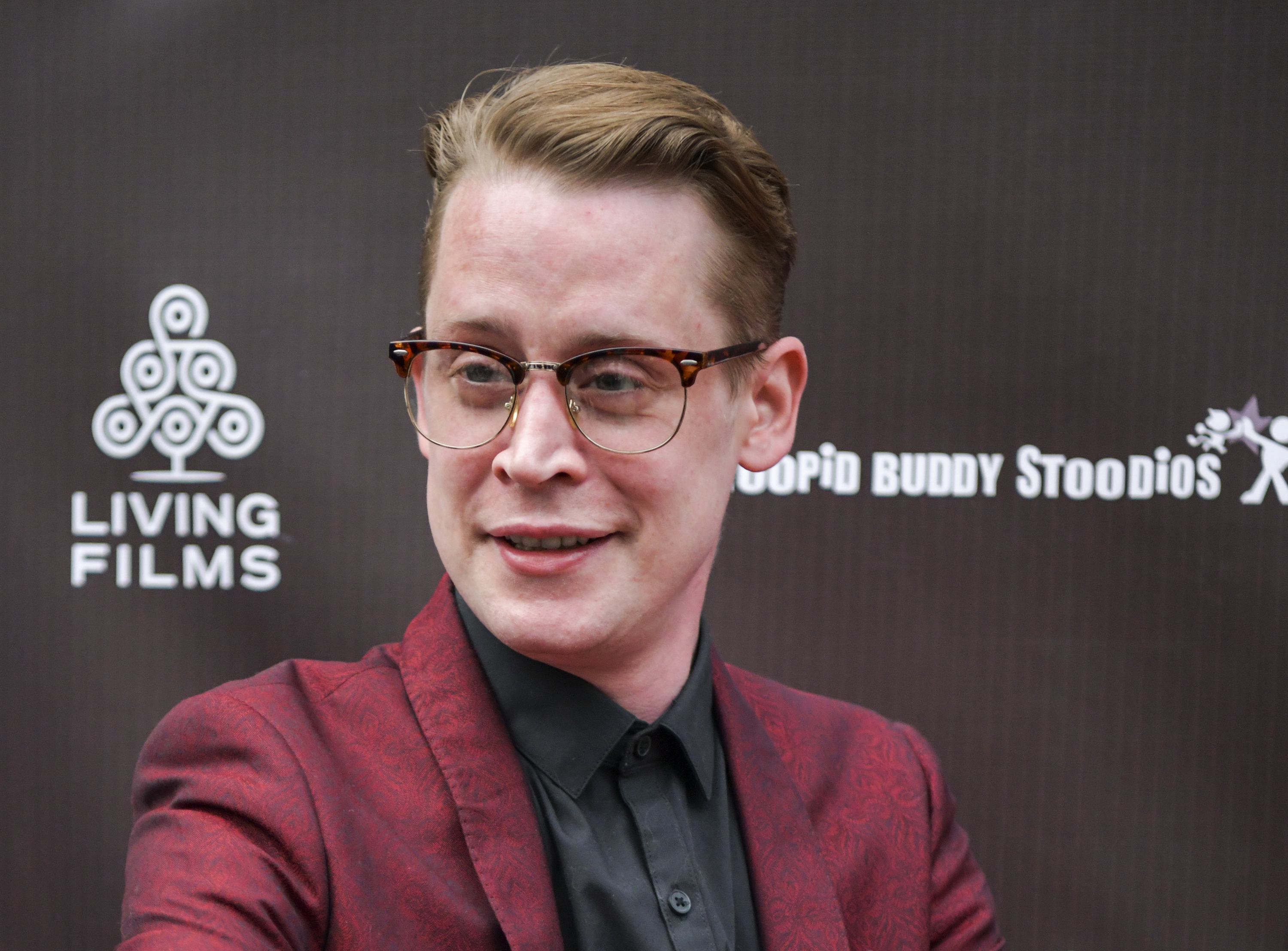 Macaulay Culkin at a red carpet event wearing a suit jacket over a dark shirt, smiling for the camera