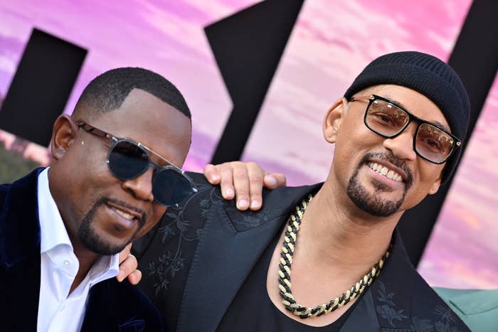 Martin Lawrence and Will Smith pose together at an event; Martin wears a dark blazer and sunglasses, Will wears a beanie, gold chain, and glasses