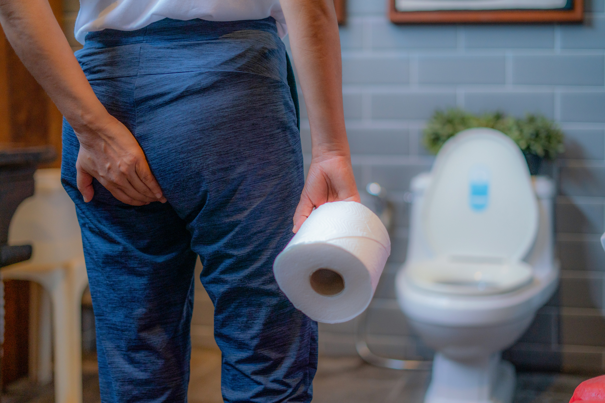Person in casual pants holding toilet paper and touching their lower back, standing in a bathroom facing a toilet