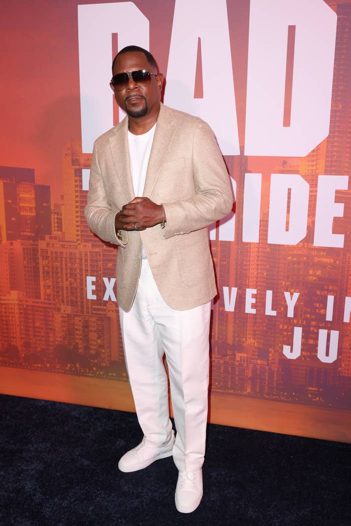 Martin Lawrence stands on a red carpet wearing a blazer, light pants, sneakers, and sunglasses