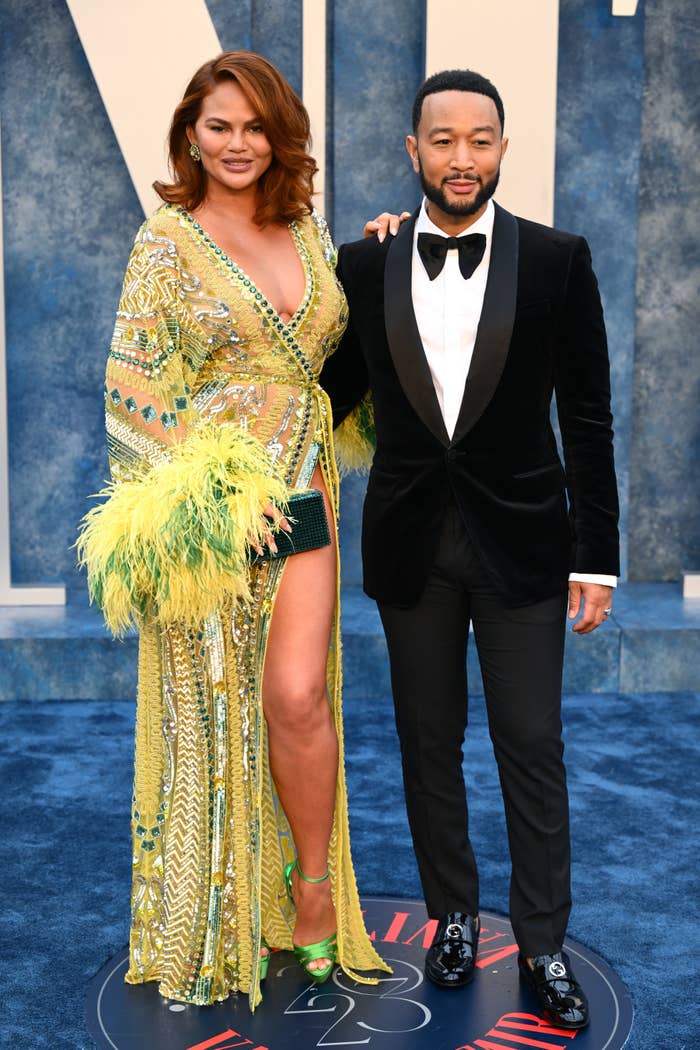 Chrissy Teigen and John Legend are on a red carpet. Chrissy wears a patterned gown with feather details and a high slit. John wears a tuxedo