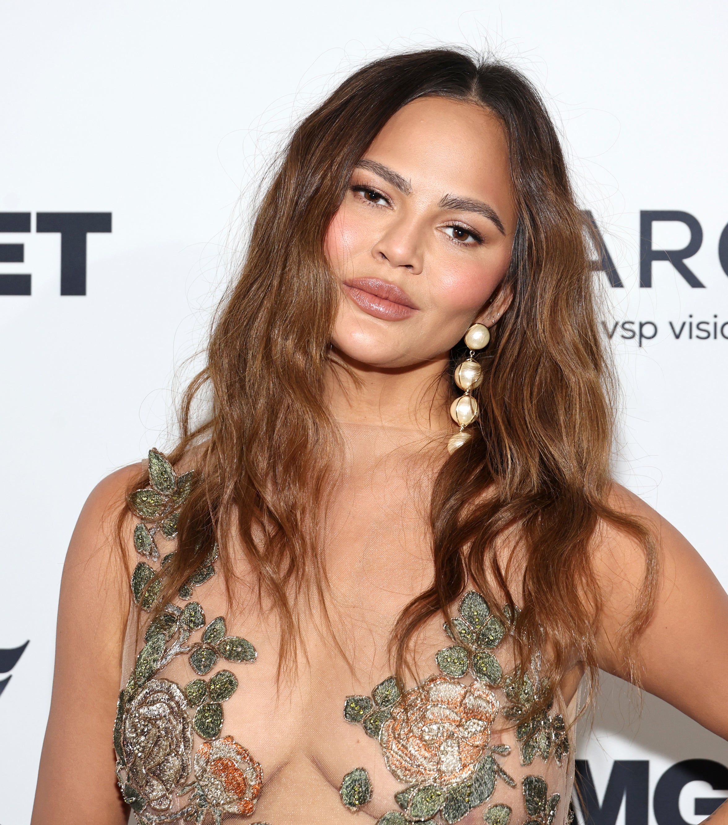 Chrissy Teigen at an event, wearing a floral embellished dress with sheer detailing, and large pearl earrings