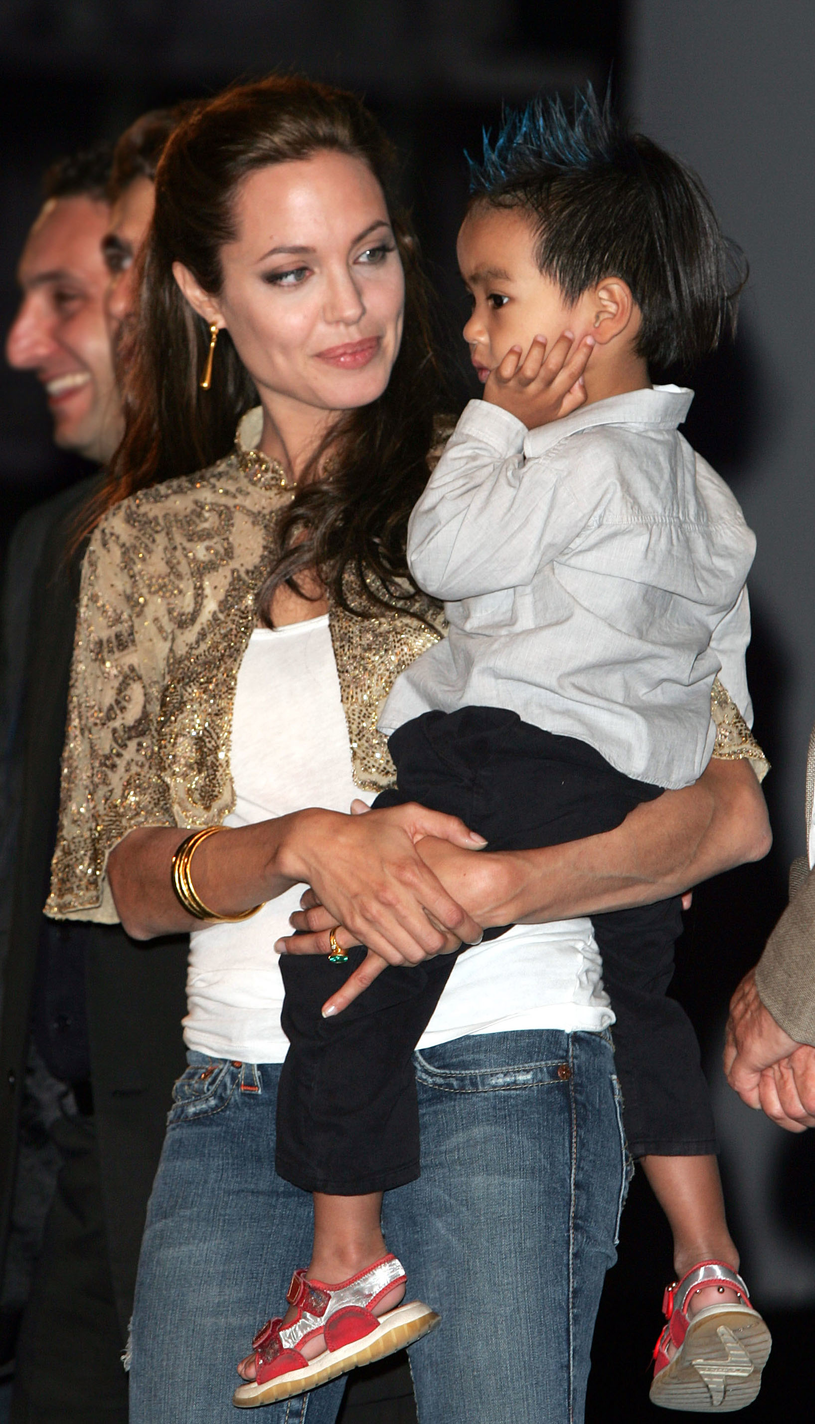 Angelina Jolie holds her young son Maddox. She wears jeans and a patterned jacket, while Maddox is dressed in a light shirt, dark pants, and sandals