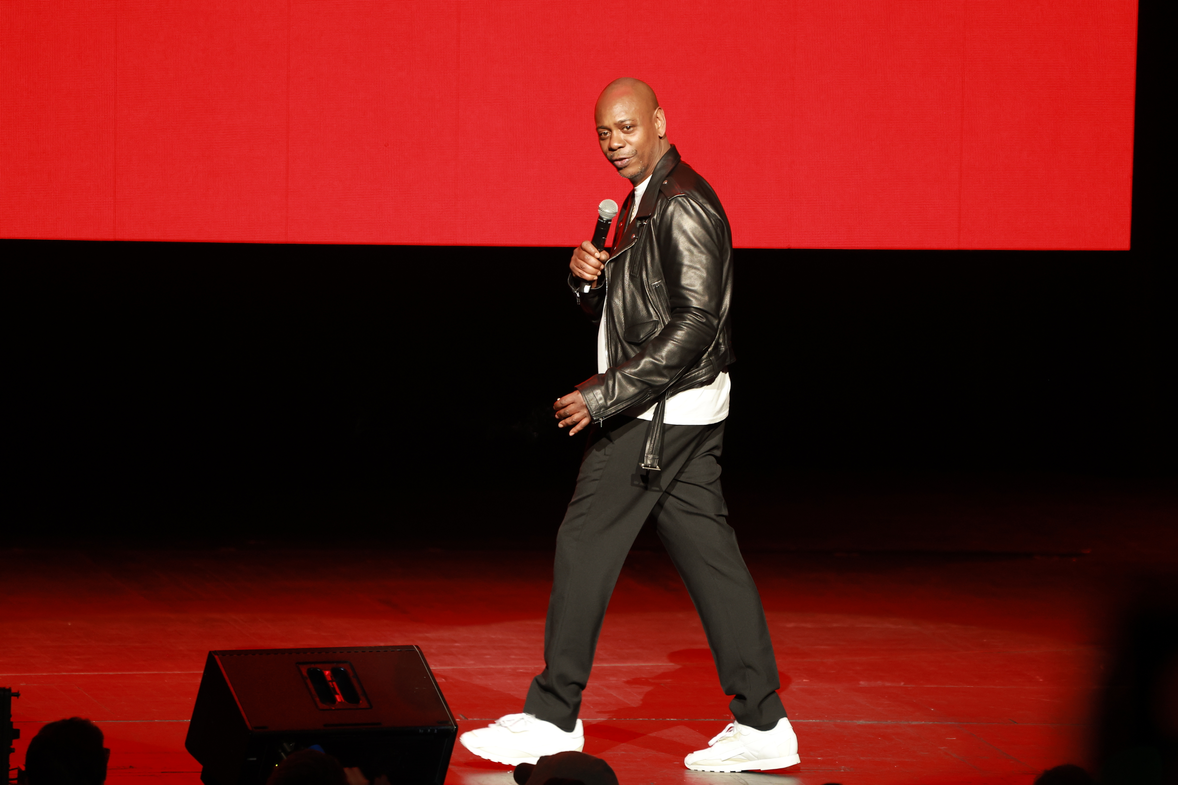 Dave Chappelle performs on stage wearing a leather jacket, white sneakers, and holding a red microphone