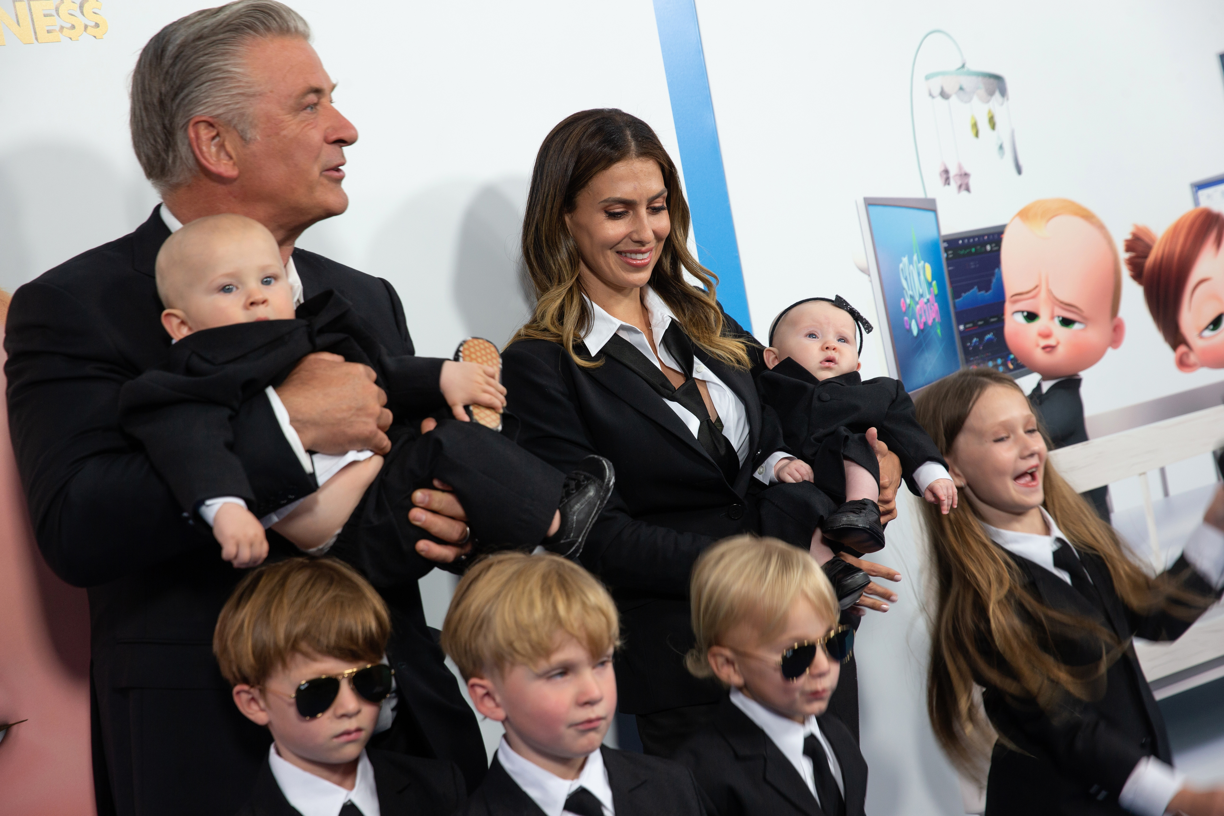 Alec Baldwin and Hilaria Baldwin pose on the red carpet with their children, all dressed in black suits and ties, at an event