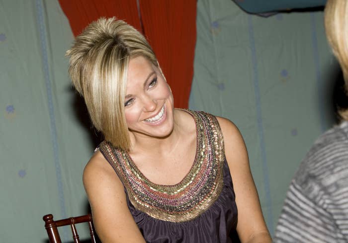 Kate Gosselin smiles seated at a table, wearing a dress with an decorated neckline and talking to an unseen person