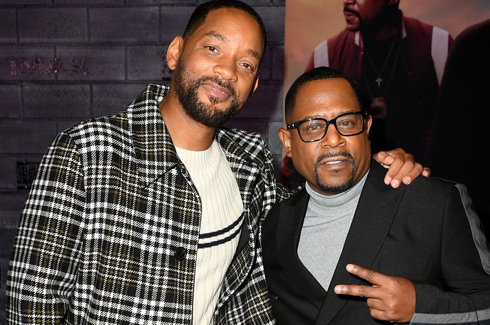 Will Smith and Martin Lawrence posing at an event. Will is wearing a checkered coat over a sweater, and Martin is wearing a black blazer with a gray turtleneck