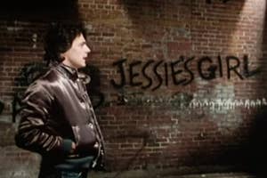 Rick Springfield stands against a brick wall with "Jessie's Girl" graffiti