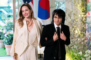Angelina Jolie in a flowing dress and trench coat with gold accessories, standing beside a young man in a black suit with a white shirt and black tie