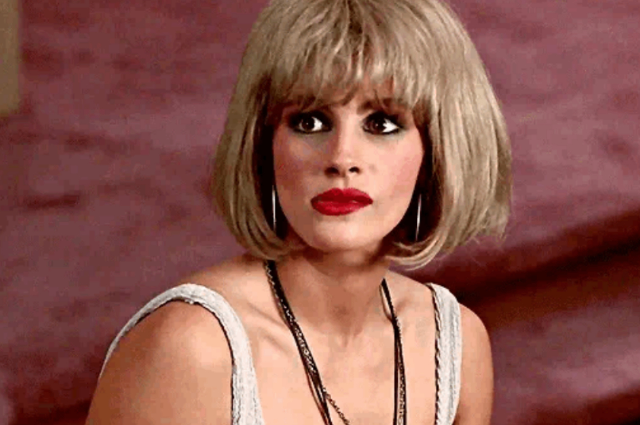 Julia Roberts, styled with a short blonde wig and bold makeup, gazes upward while wearing a sleeveless top and layered necklaces. The scene hints at a dramatic moment