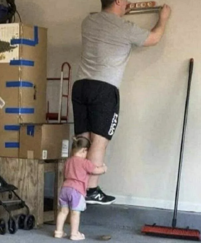 Adult wearing a grey t-shirt and black shorts uses a level on a wall while a child holds onto their legs in a room with moving boxes and a broom nearby