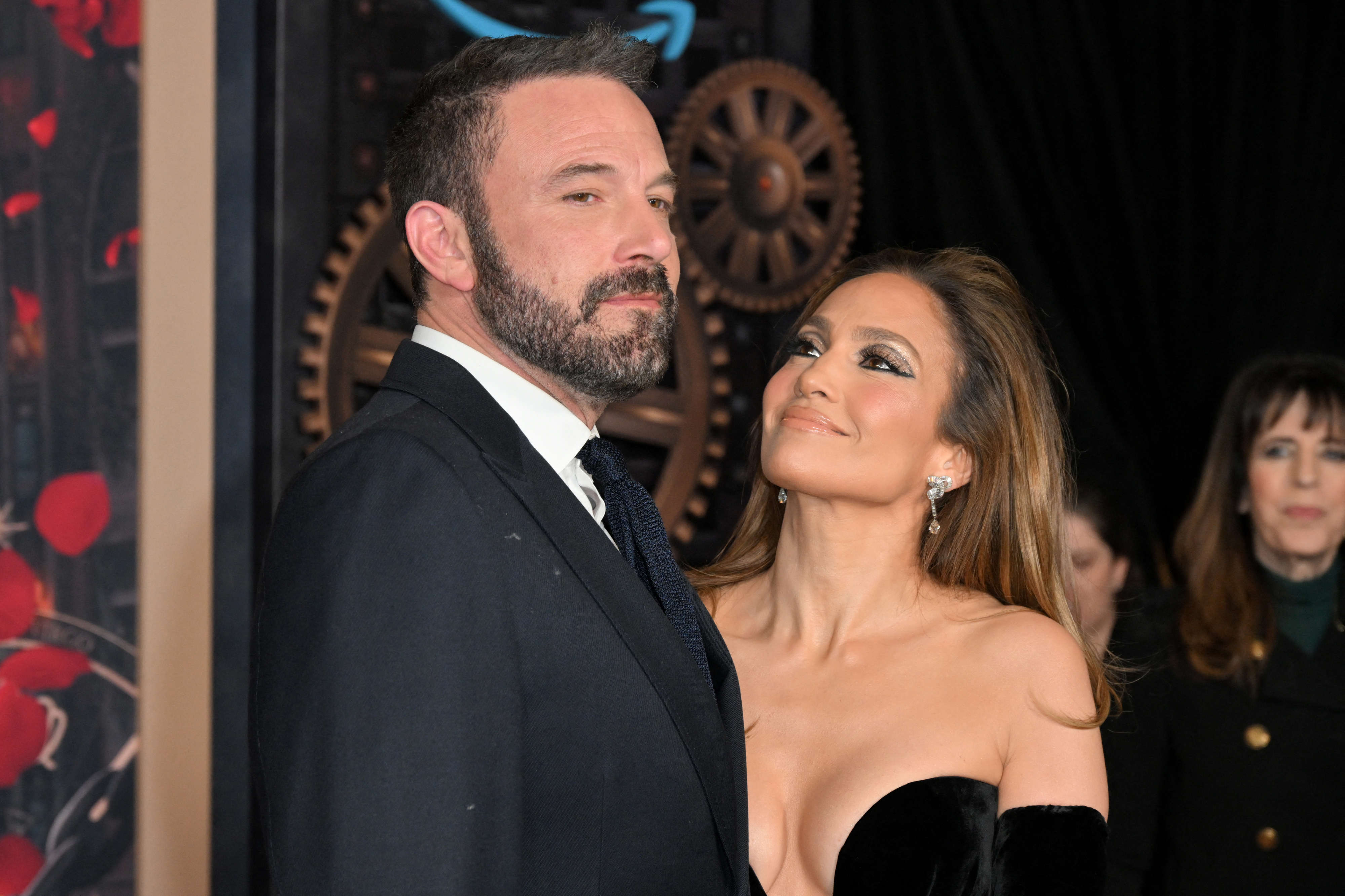 Ben Affleck in a suit and Jennifer Lopez in an elegant off-the-shoulder gown standing together, with Jennifer looking at Ben affectionately