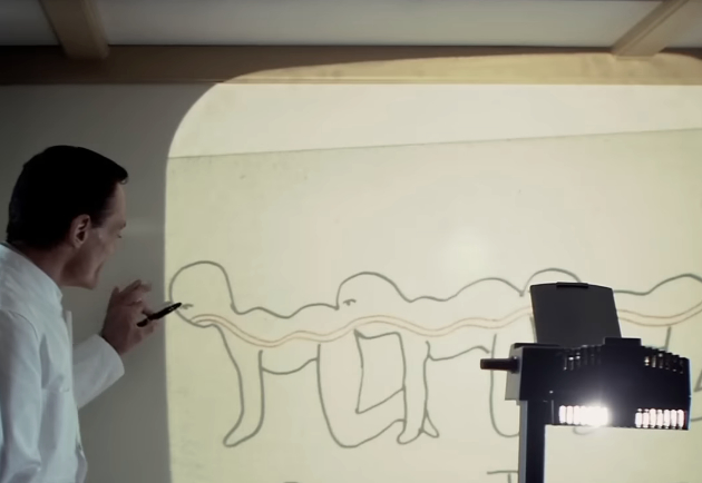 A person is pointing at a diagram showing a drawing of three human figures connected in a sequence, projected on a screen