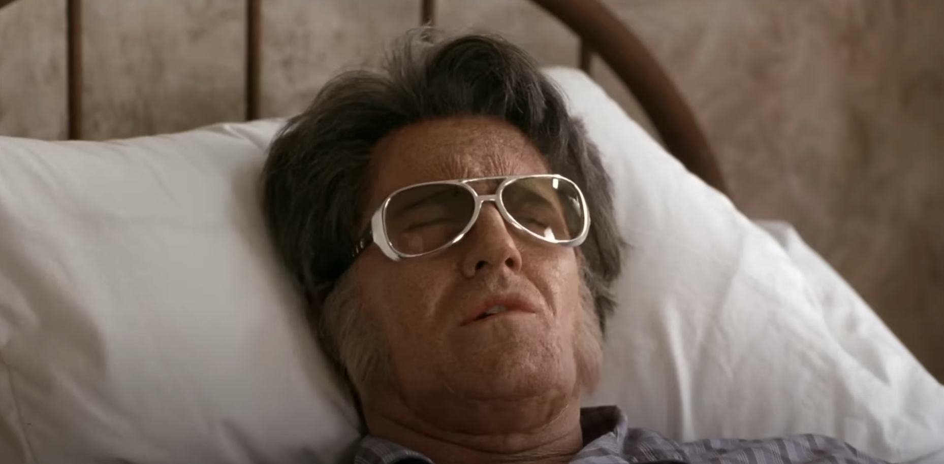 Elvis with gray hair and large glasses is lying in bed with his eyes closed