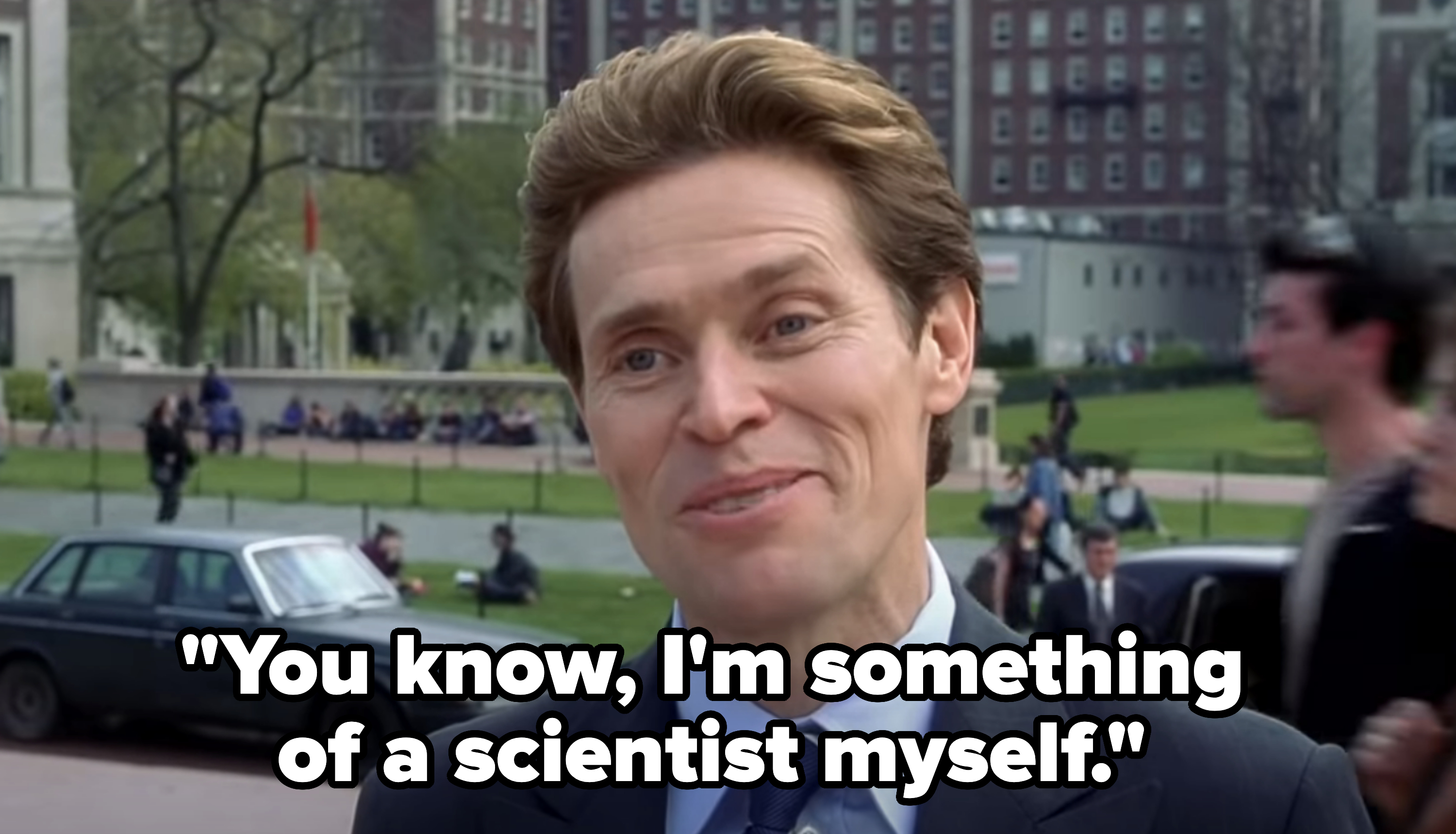 Willem Dafoe in a suit and tie, smiling outdoors with buildings and people in the background