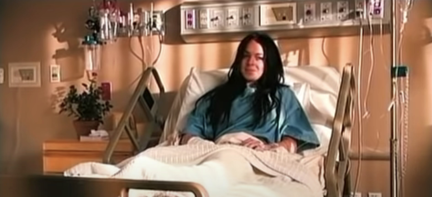 Lindsay Lohan lying in a hospital bed in a medical setting, wearing a hospital gown with medical equipment visible in the background