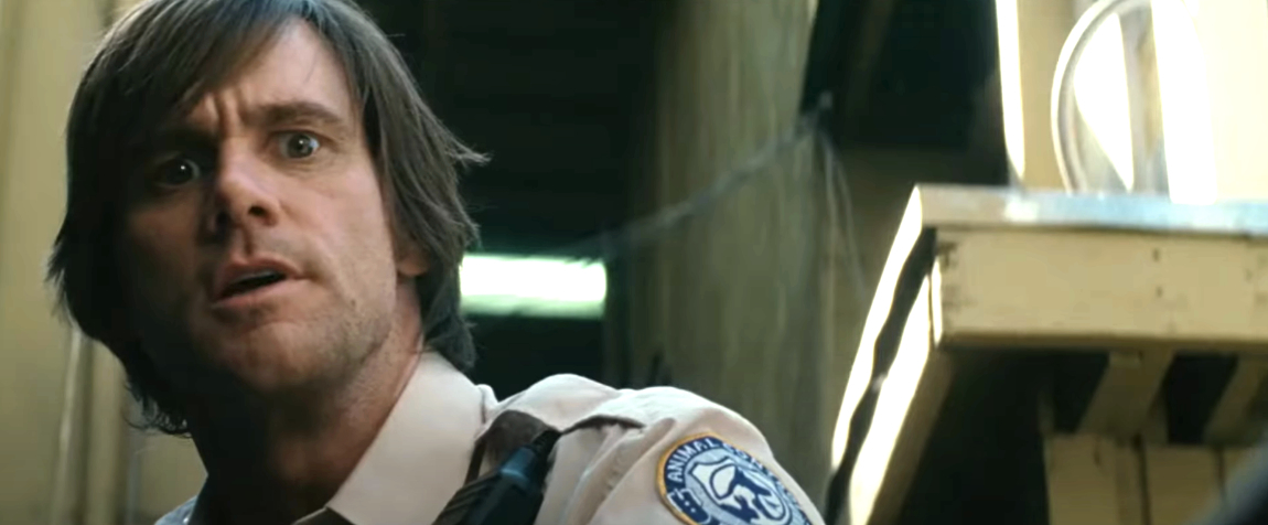 Jim Carrey, wearing a beige security guard uniform with a patch on the sleeve, appears surprised in a dimly lit industrial setting