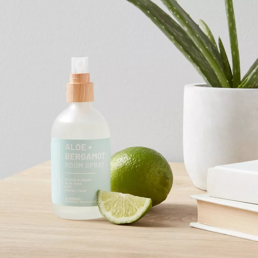 A bottle of Aloe + Bergamot room spray stands on a wooden surface beside a lime and a sliced lime wedge, with a potted plant and book in the background