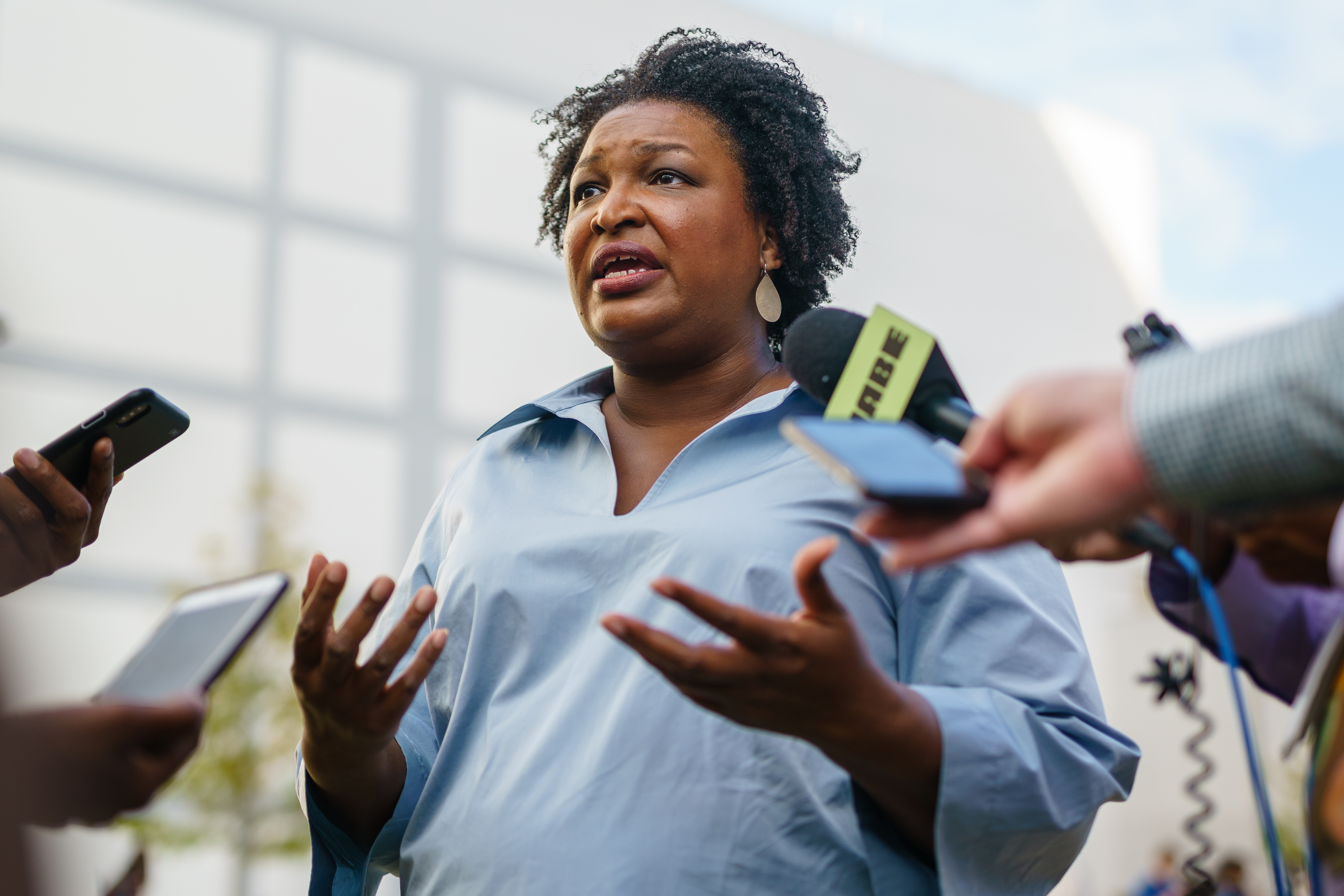 Stacey Abrams speaks to the press, wearing a light blouse, gesturing with her hands, surrounded by microphones and recording devices