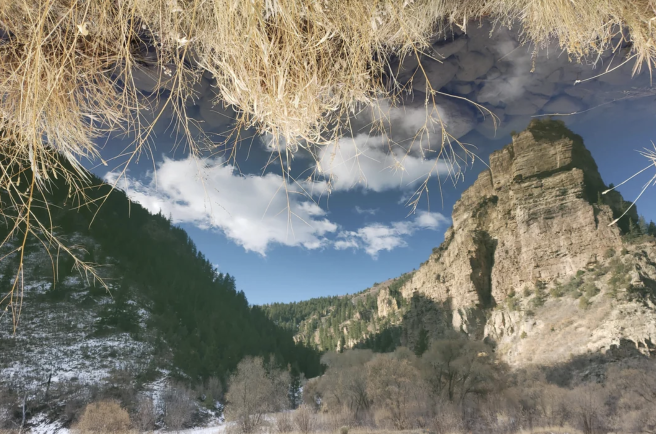 Reflection of mountain and sky in clear water creates an upside-down landscape illusion with dry grass, clouds, and steep cliffs